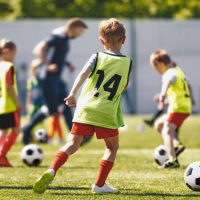 School Children Play Soccer Football Training Game With Coach. Kids Kicking Sports Balls on Grass Field. Football Practice For Youth